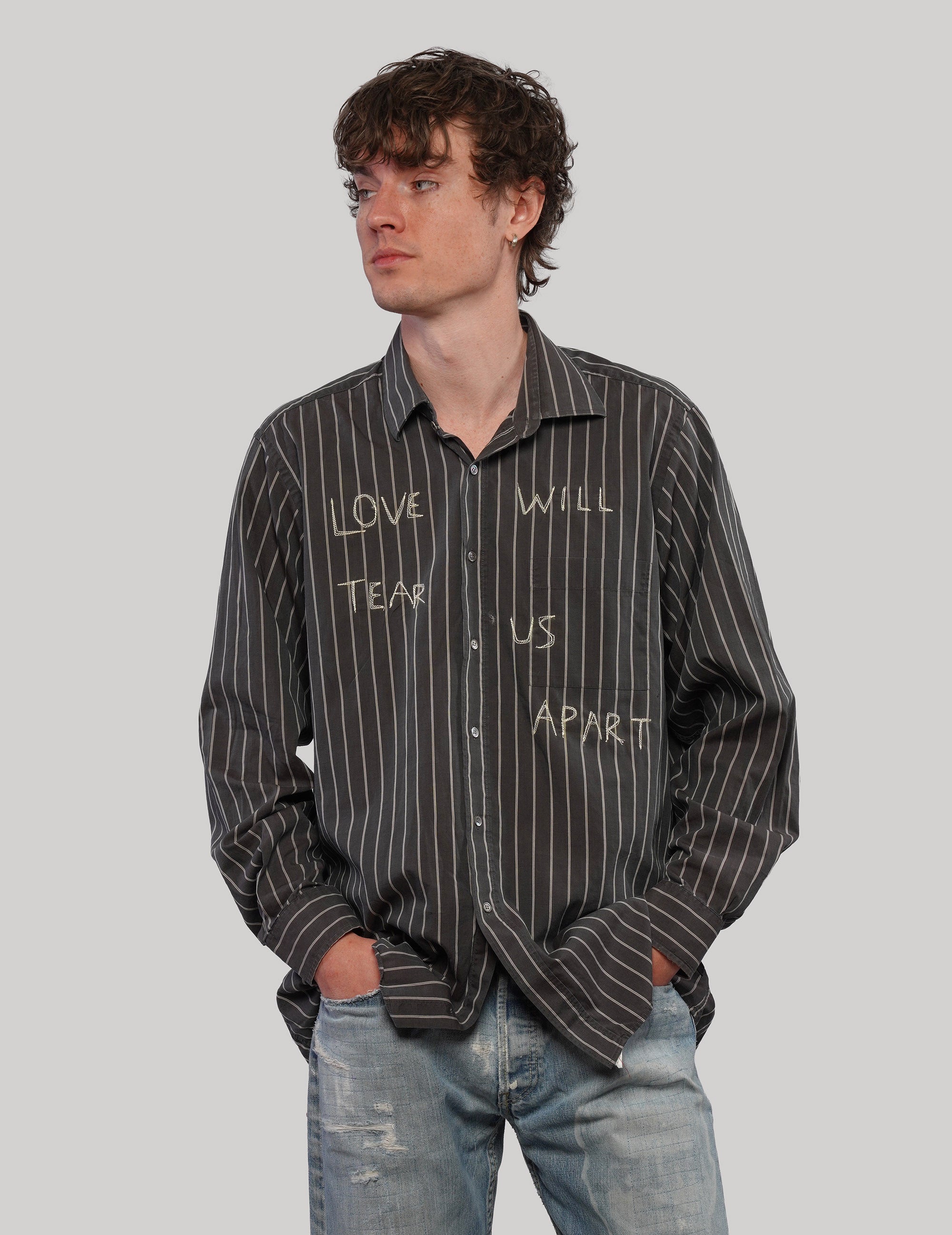 Model wearing a black 'Love Will' pinstripe shirt with hands in jeans pockets, showing the front graphic lettering.