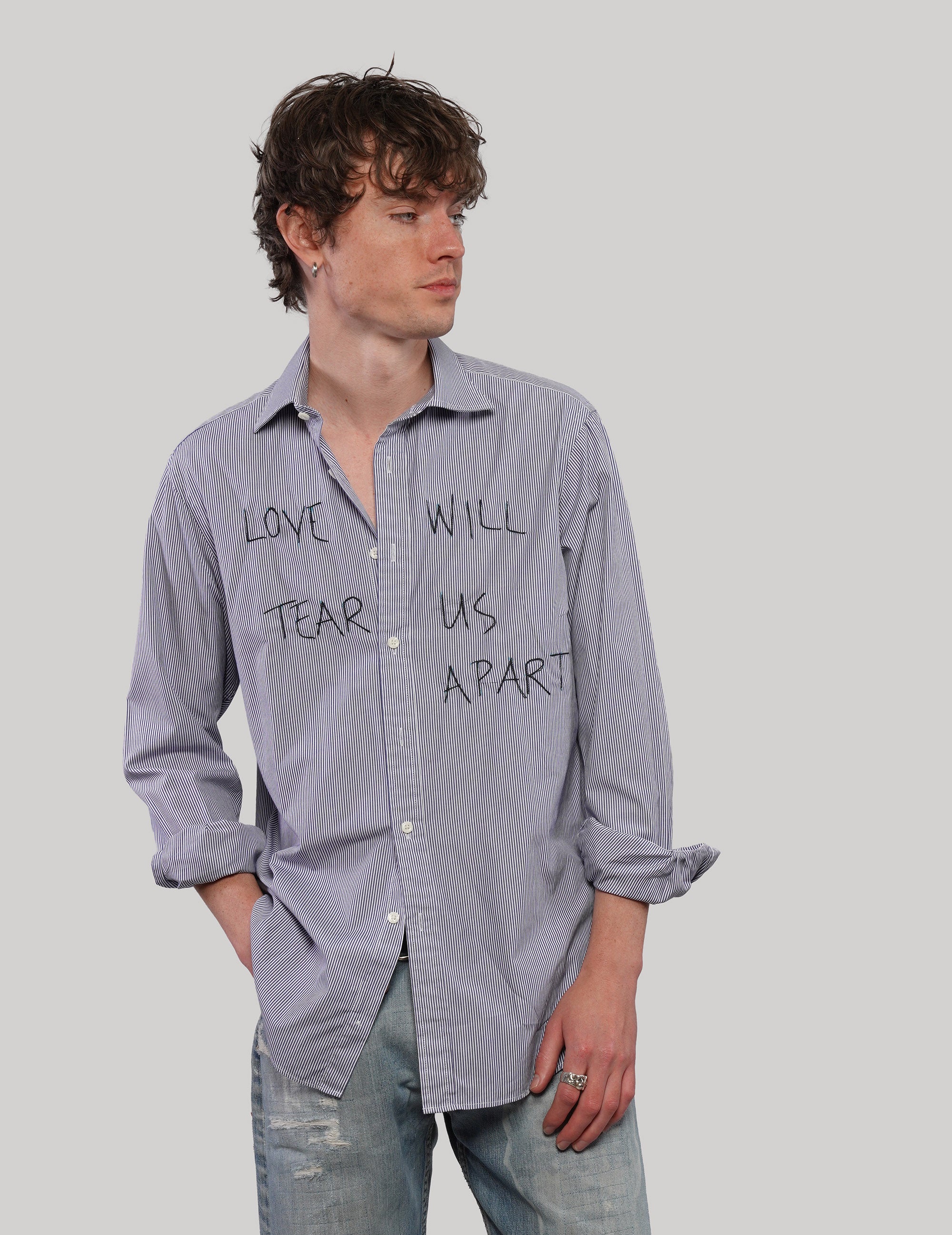 Model wearing a blue pinstripe 'Love Will' shirt with black graphic lettering, looking to the side with one hand in his jeans pocket.