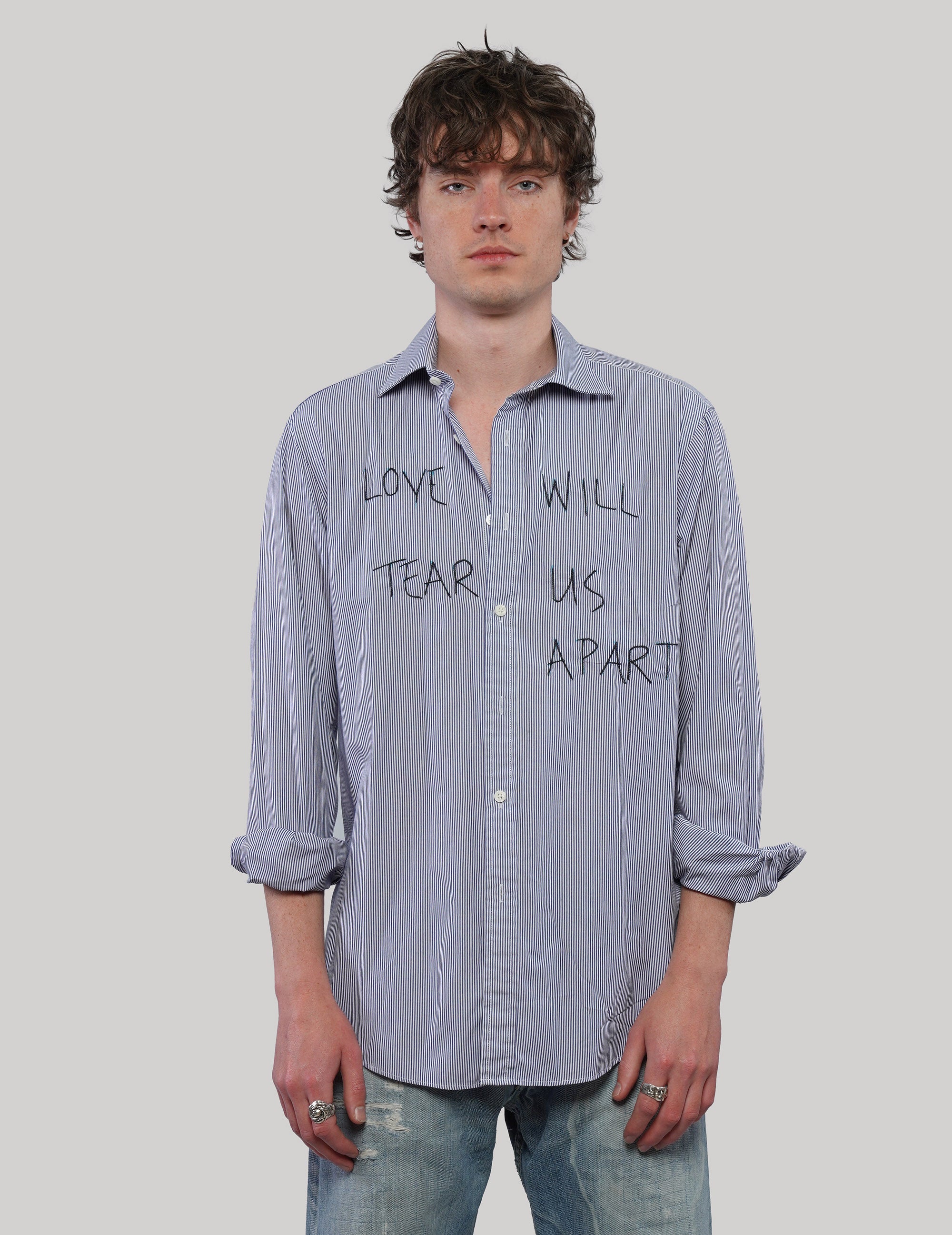 Model wearing a blue and white pinstripe 'Love Will' cotton shirt with black handwritten-style lettering and rolled sleeves.