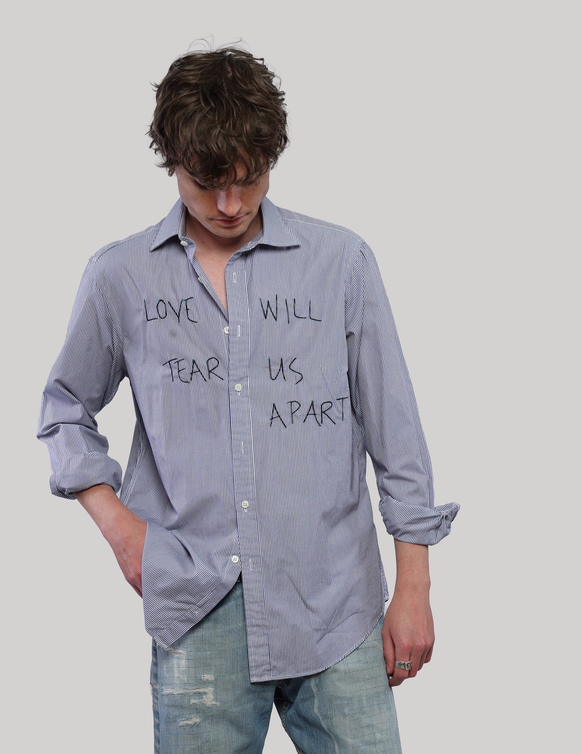 Model looking down wearing a blue striped 'Love Will' shirt with black front lettering, rolled sleeves and distressed jeans.