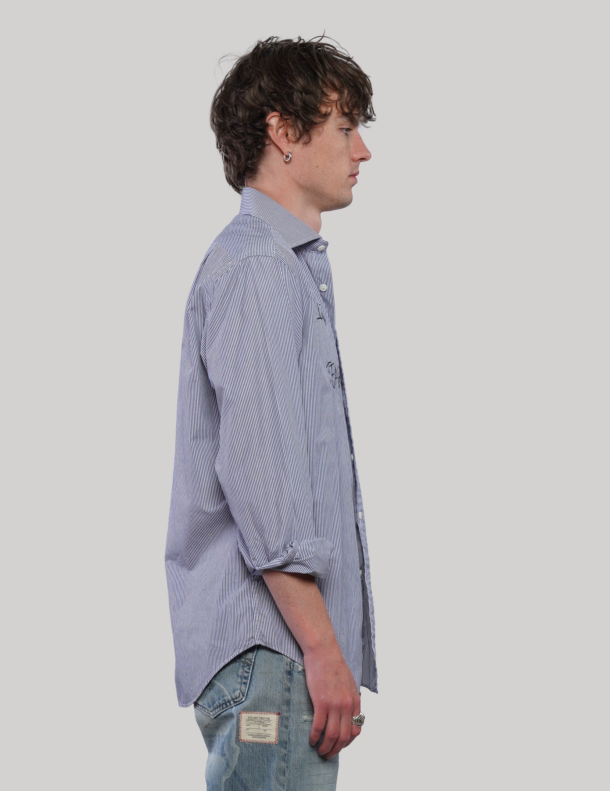 Side view of a blue and white vertically striped cotton shirt showing its loose silhouette, long sleeves and curved hem.
