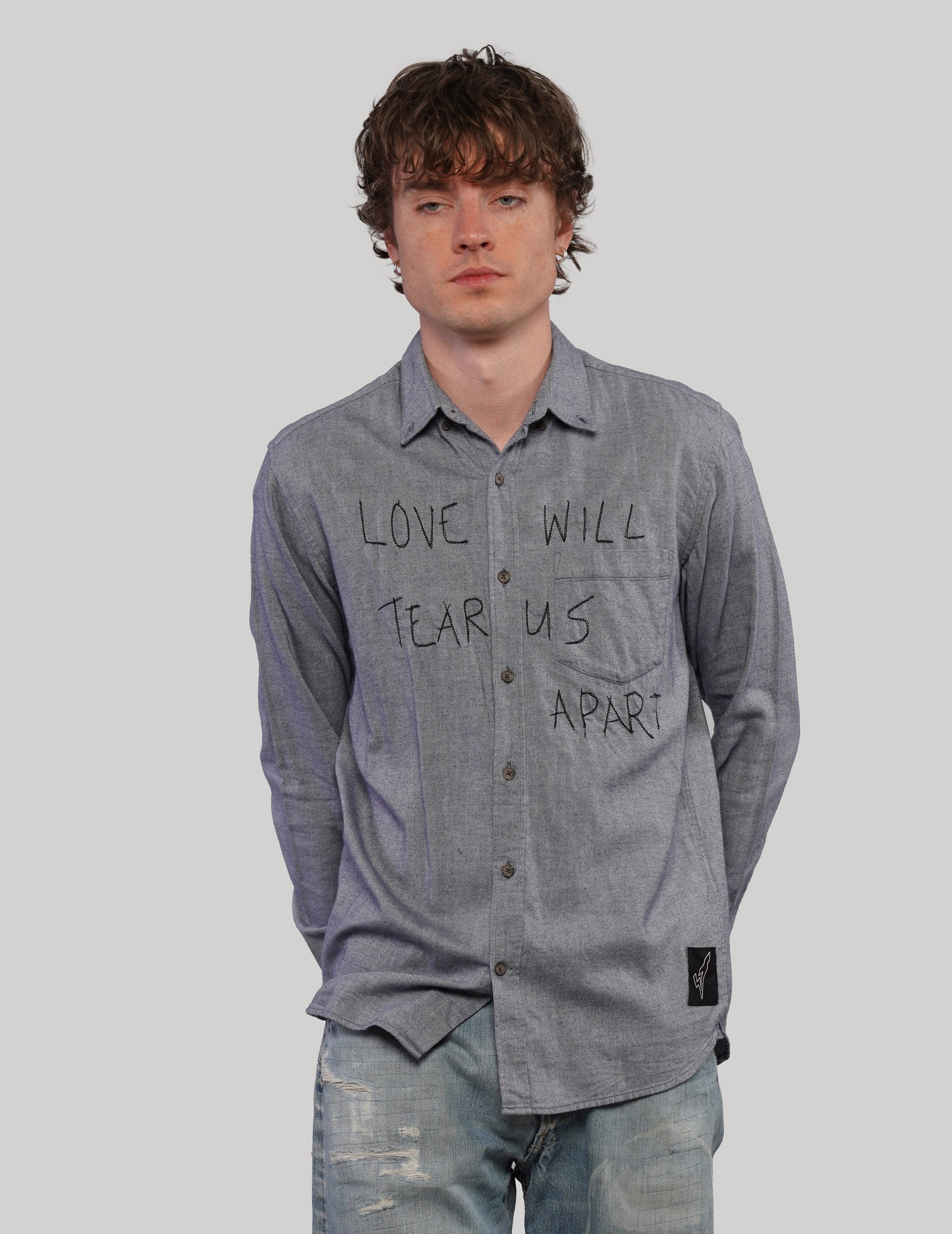 Model wearing a grey 'Love Will' cotton shirt with black handwritten-style lettering, pointed collar and dark buttons.