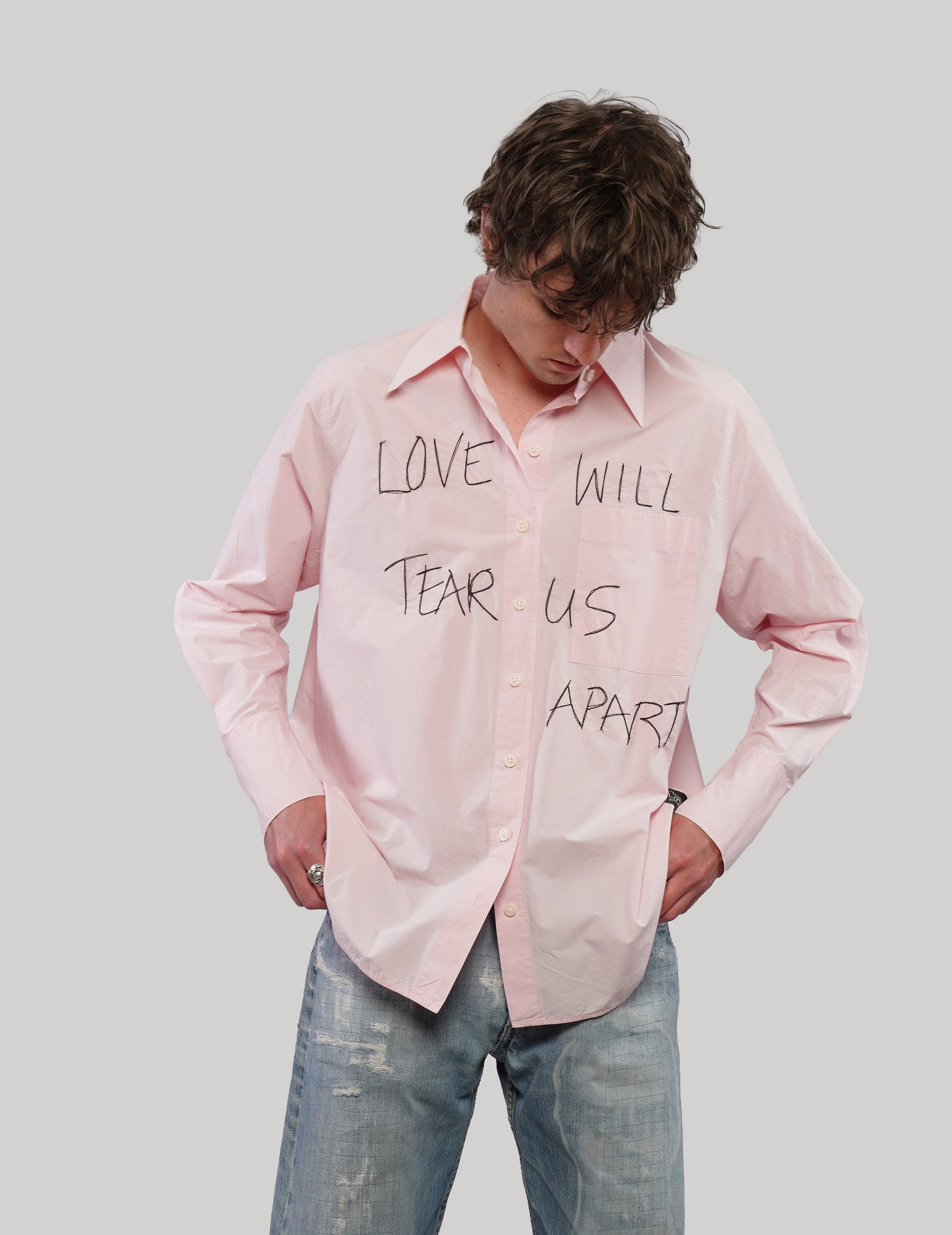 Model looking down wearing a pink 'Love Will' graphic shirt with black lettering, styled with distressed blue jeans.