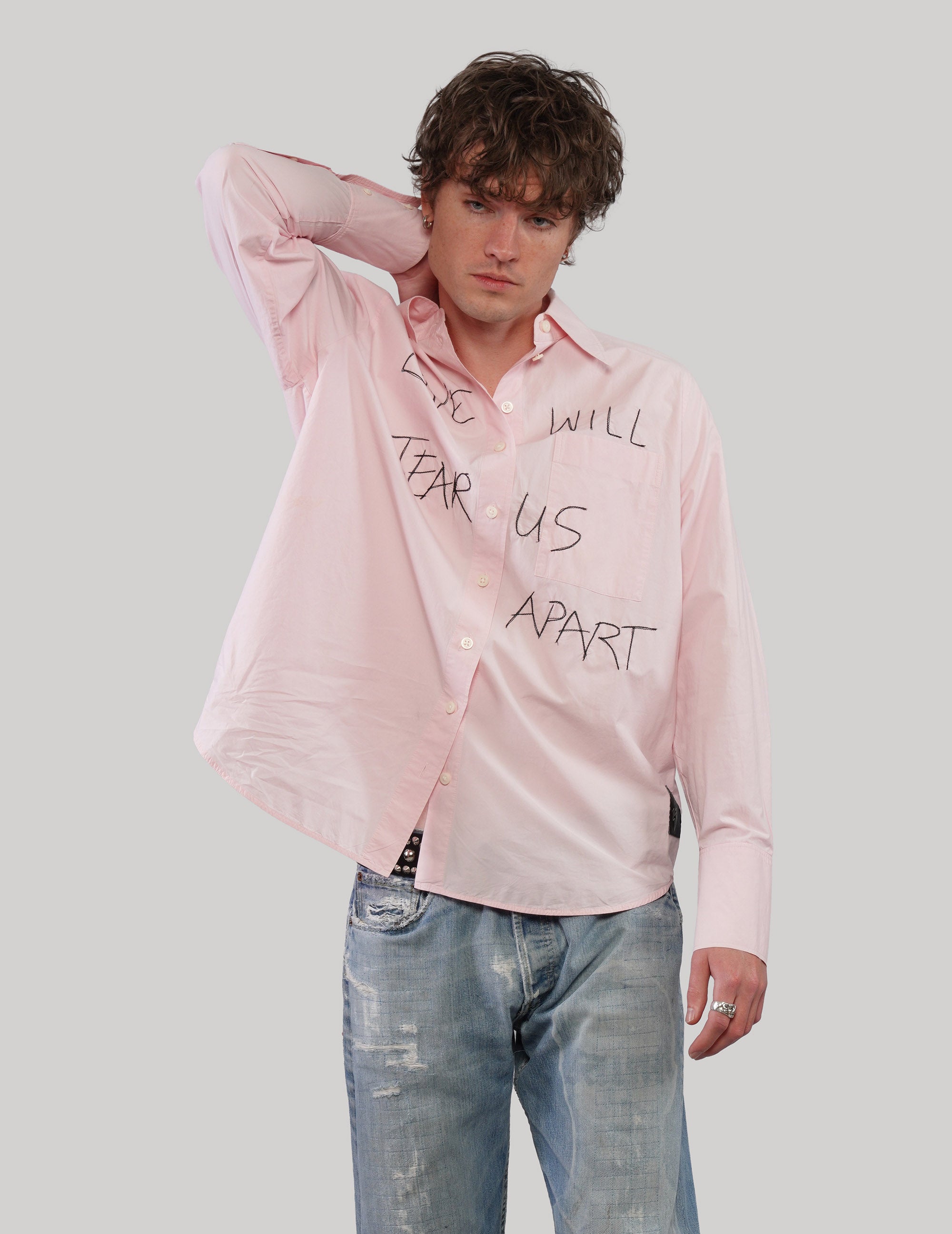 Model wearing a pink 'Love Will' cotton shirt with black graphic lettering, one hand resting behind his head.