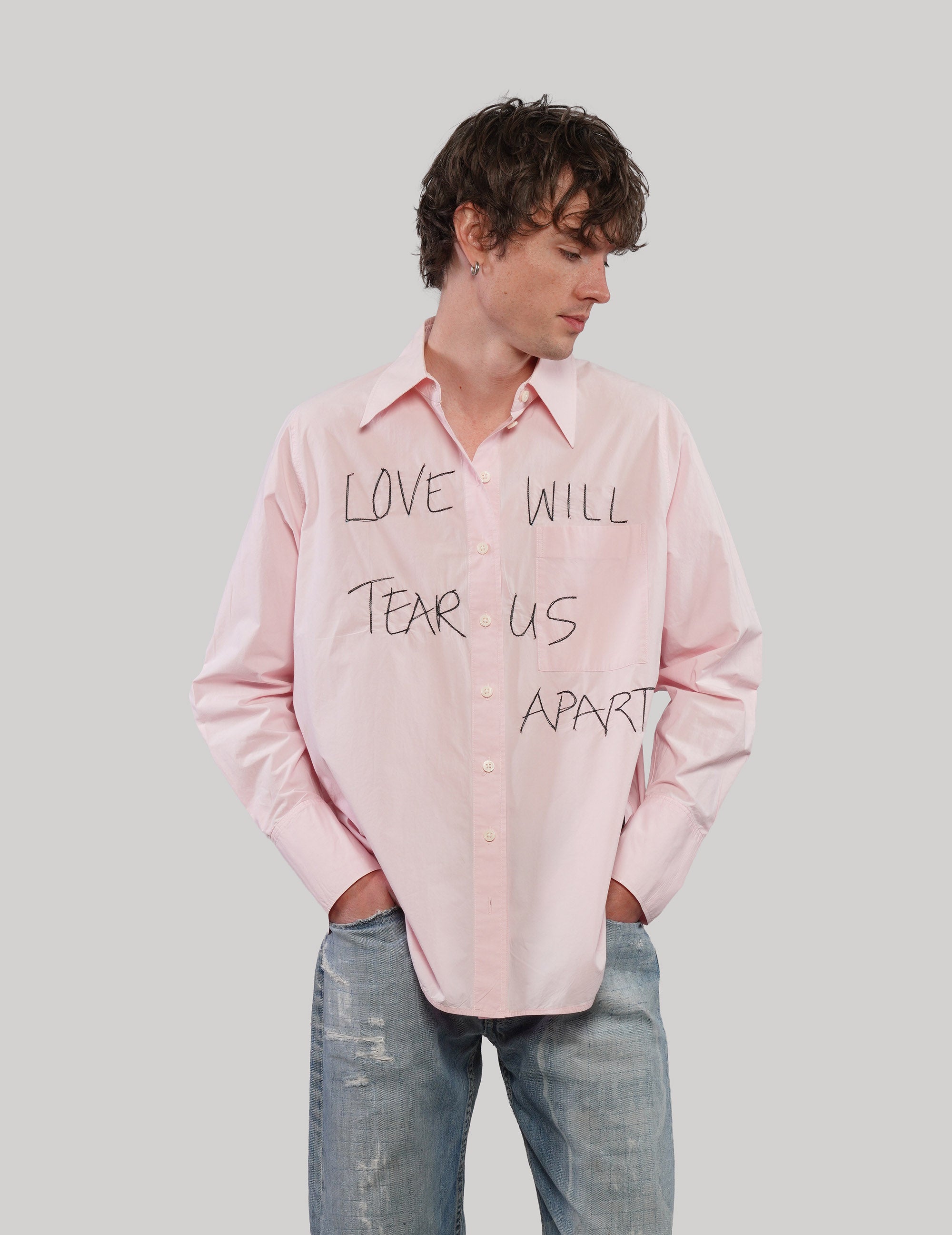 Model wearing a pink 'Love Will' cotton shirt with black front lettering, one hand in his jeans pocket.