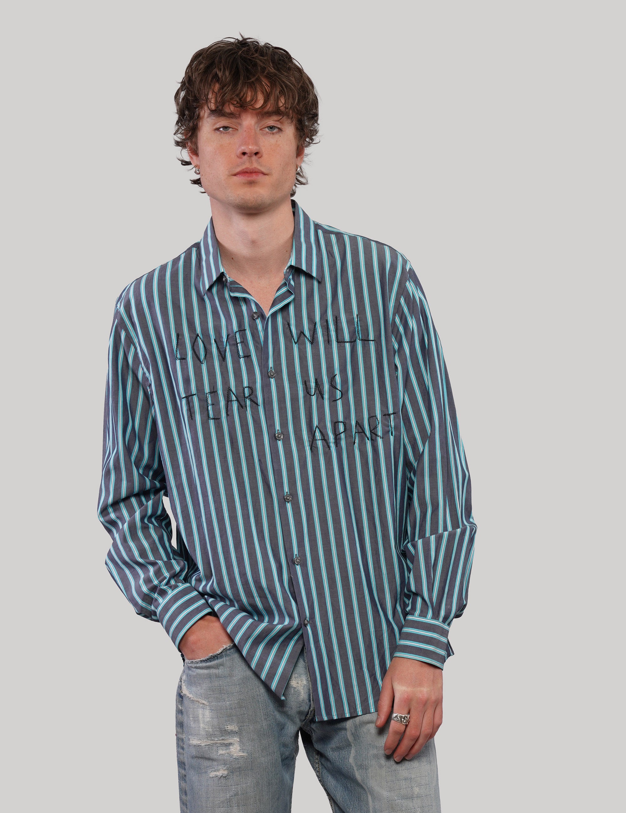 Model wearing a teal and light blue striped 'Love Will' cotton shirt with black graphic lettering and button fastening.