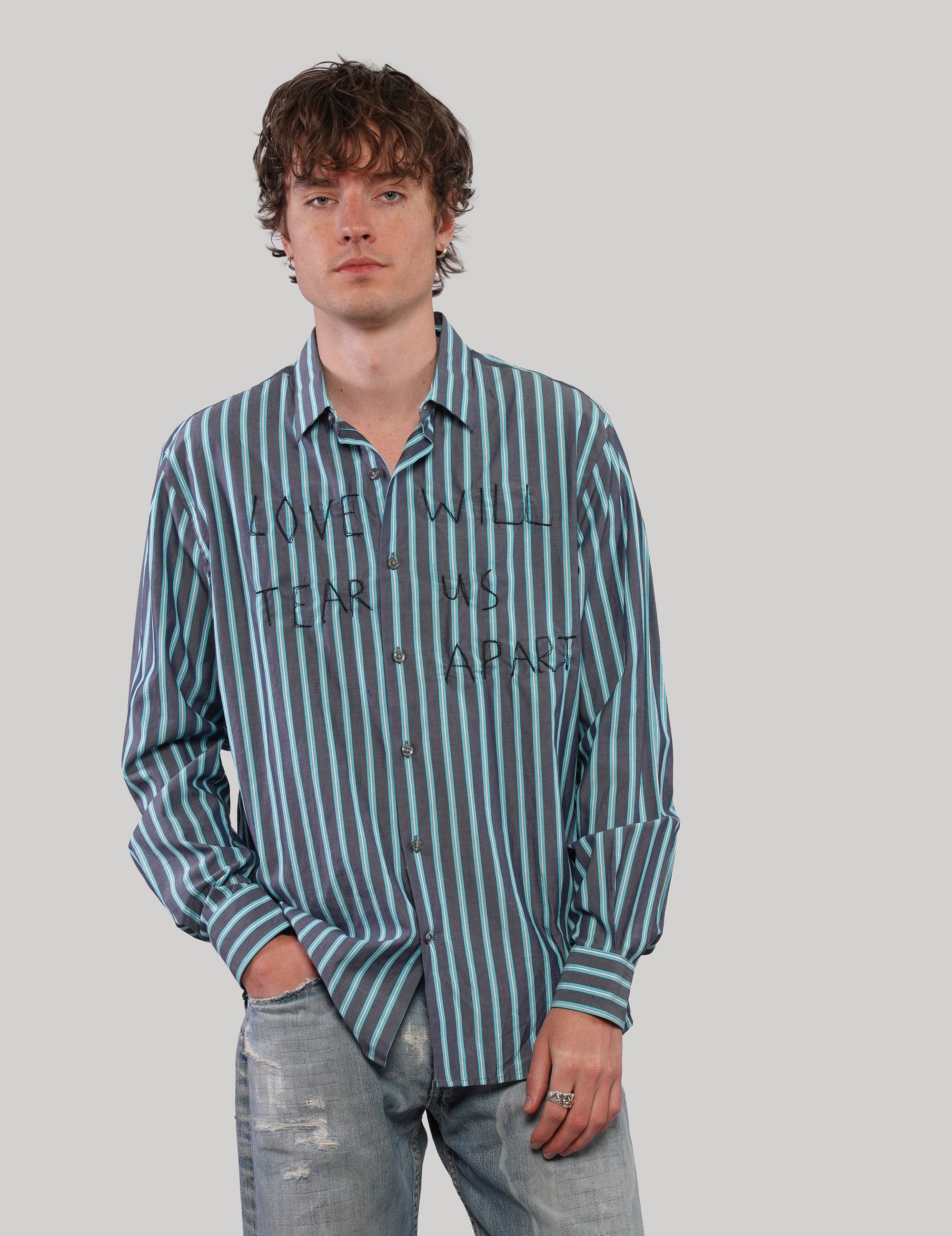 Model wearing a teal striped 'Love Will' cotton shirt with hands in pockets, styled with distressed blue jeans.
