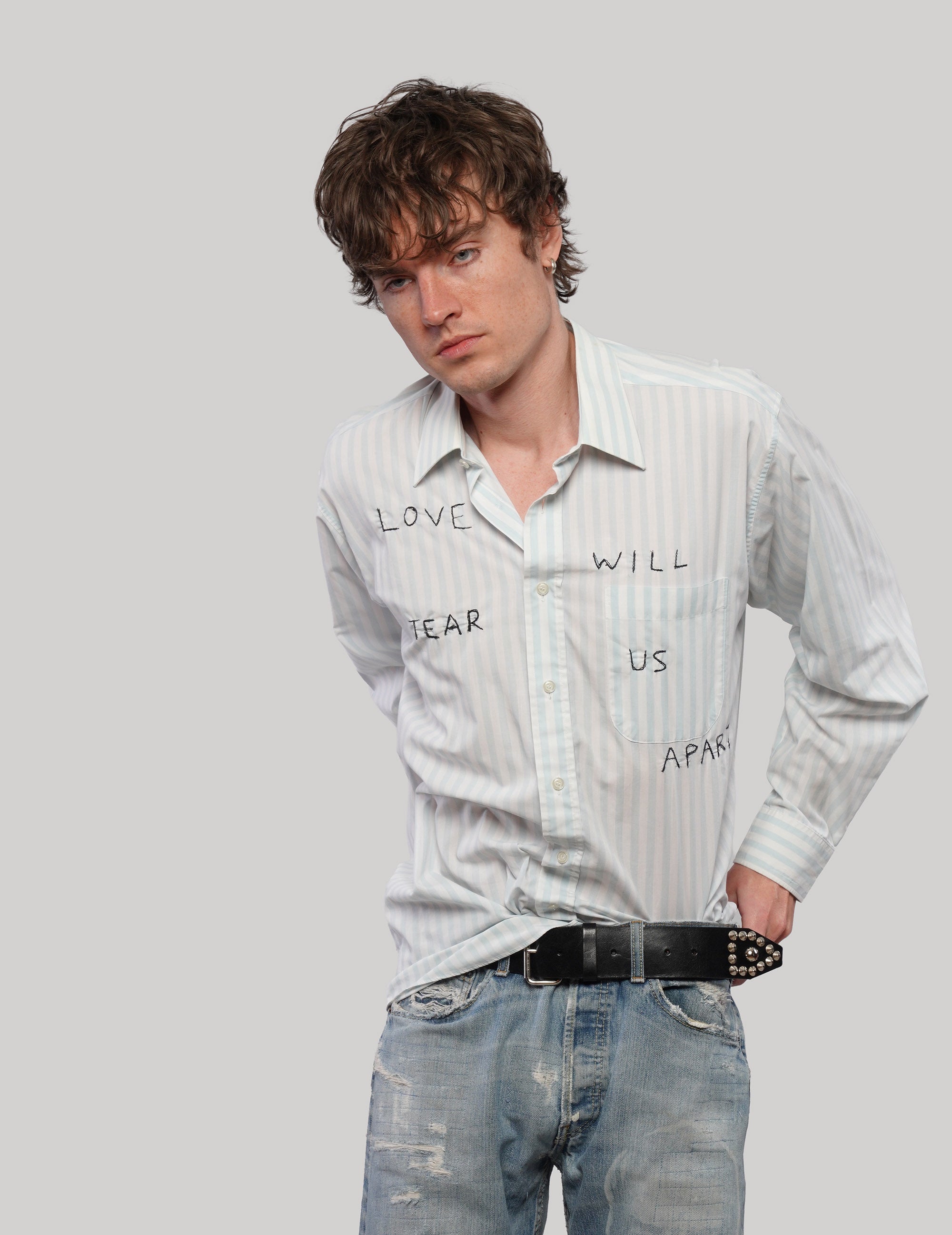 Model wearing a white 'Love Will' striped shirt with black lettering, tucked into distressed blue jeans.