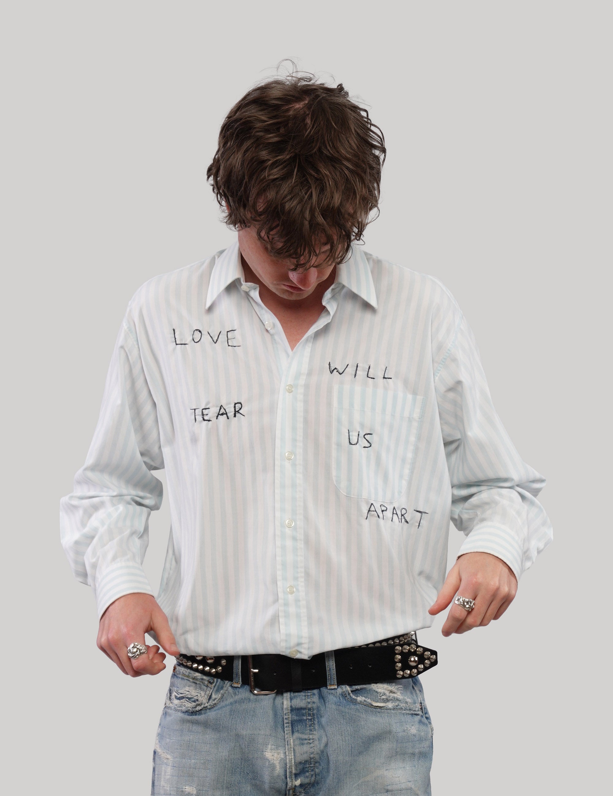 Model looking down while adjusting a white striped 'Love Will' shirt with long sleeves and graphic lettering.