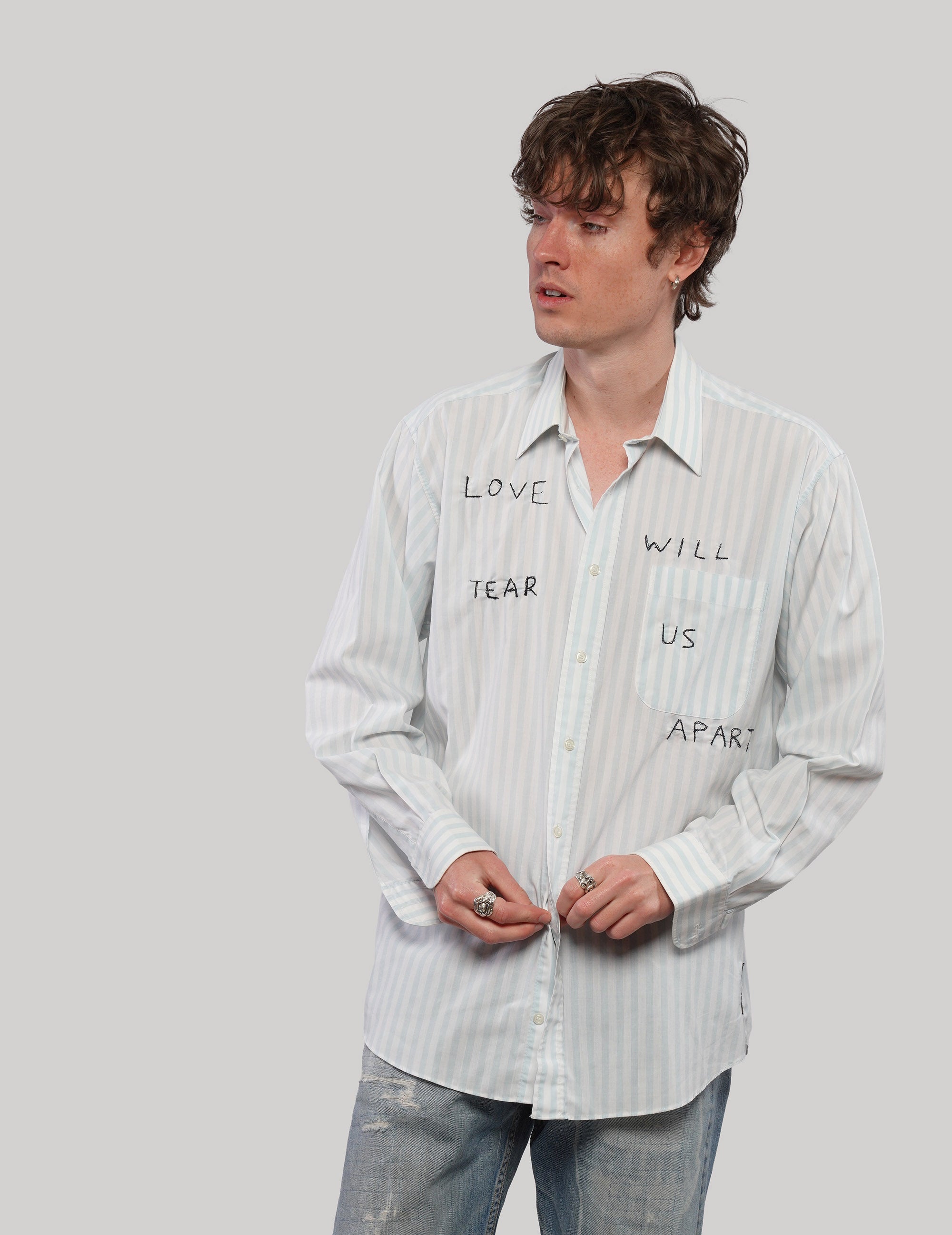 Model wearing a white striped 'Love Will' cotton shirt with pointed collar and black lettering, styled with jeans.