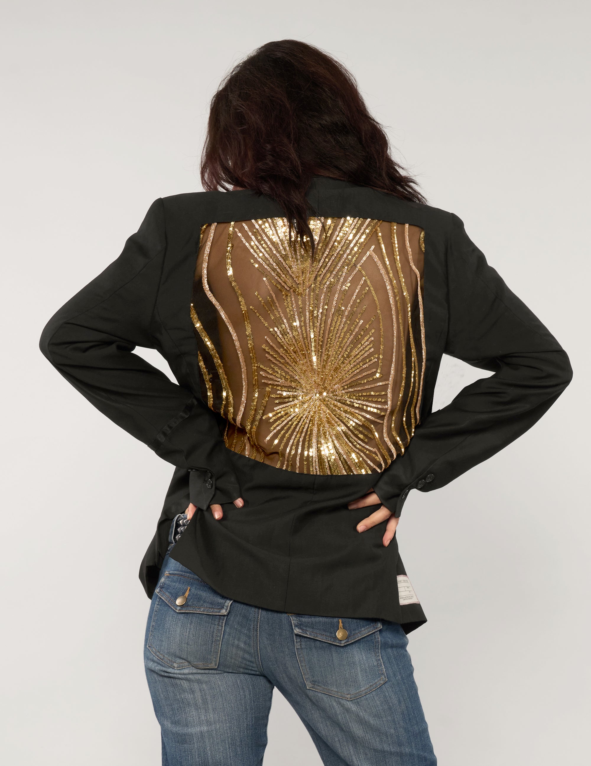 Model wearing a black vintage tuxedo jacket with gold sequin starburst back panel, hands on hips, styled with blue jeans.
