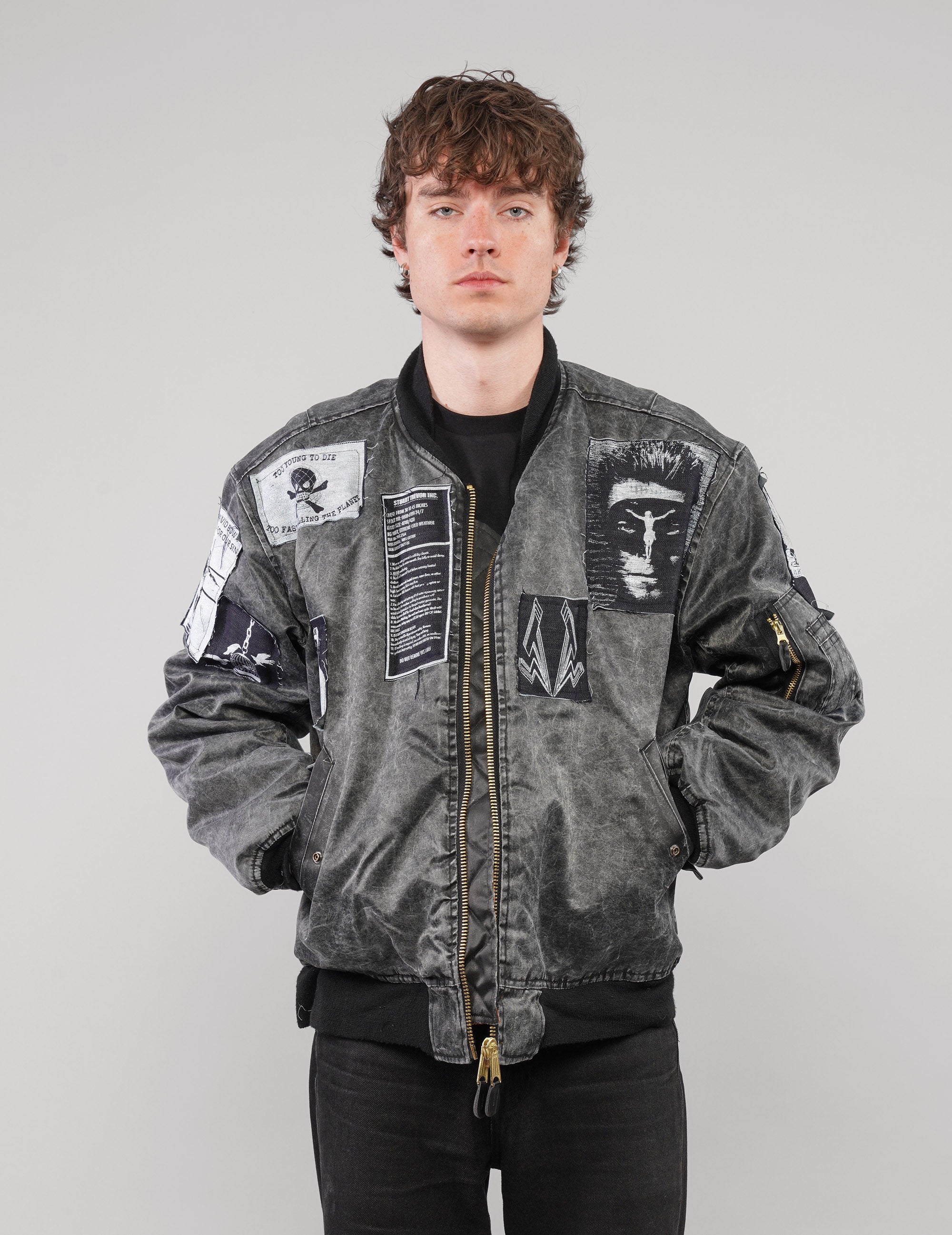 Front view of the zipped charcoal bomber jacket with a lightning chest patch, worn with hands in the pockets.