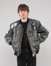 Charcoal patched bomber jacket worn unzipped over a black T-shirt, with the model’s hands in the front pockets.