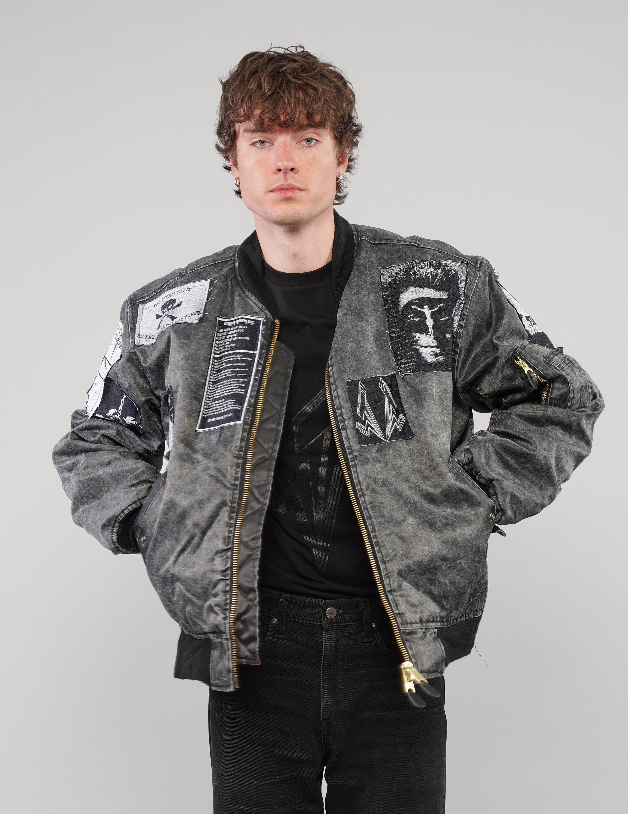 Charcoal patched bomber jacket worn unzipped over a black T-shirt, with the model’s hands in the front pockets.
