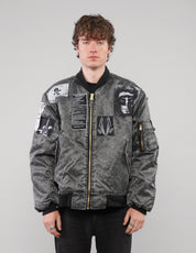 Model wearing a charcoal patched bomber jacket zipped up, showing monochrome chest and sleeve graphics.