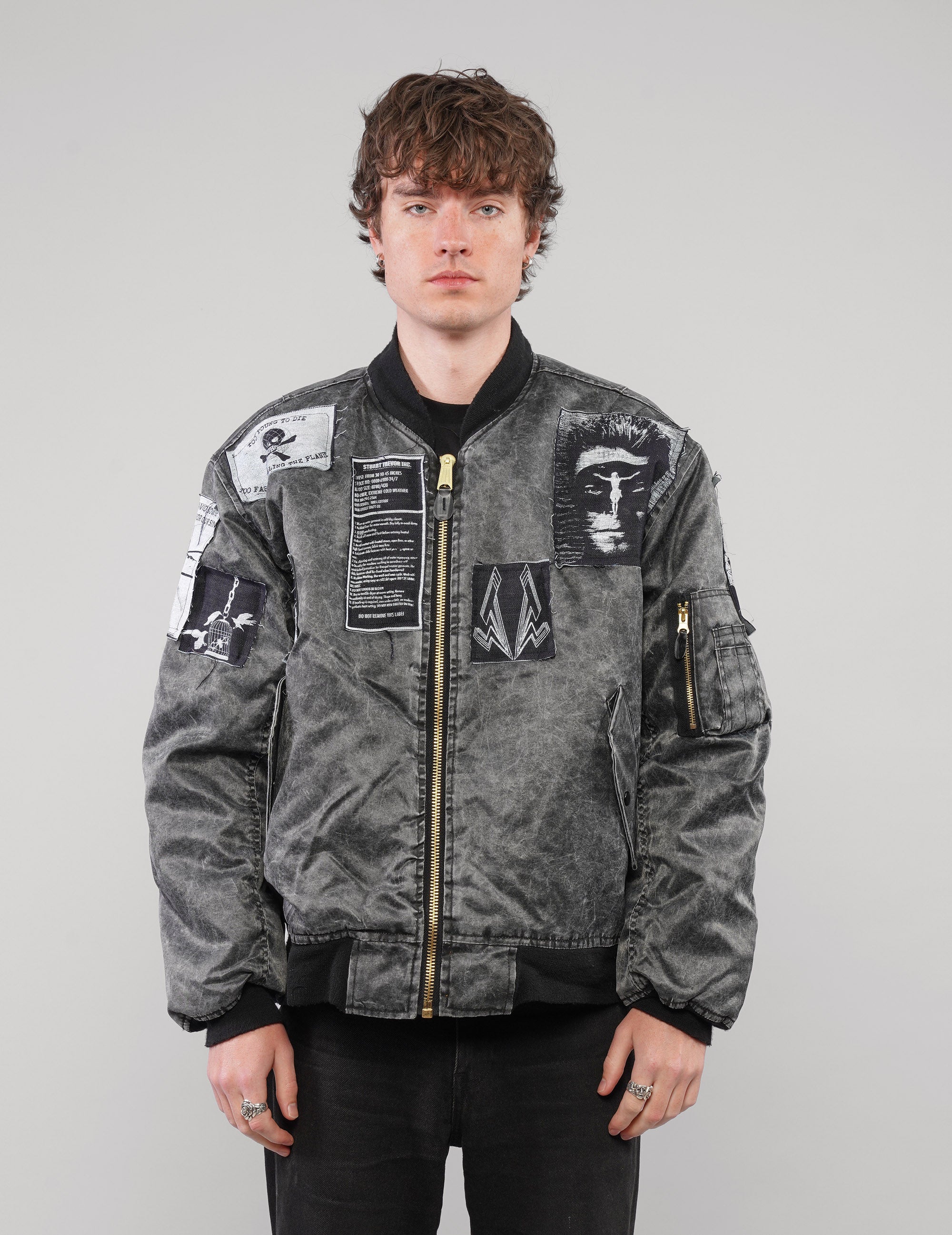Model wearing a charcoal patched bomber jacket zipped up, showing monochrome chest and sleeve graphics.