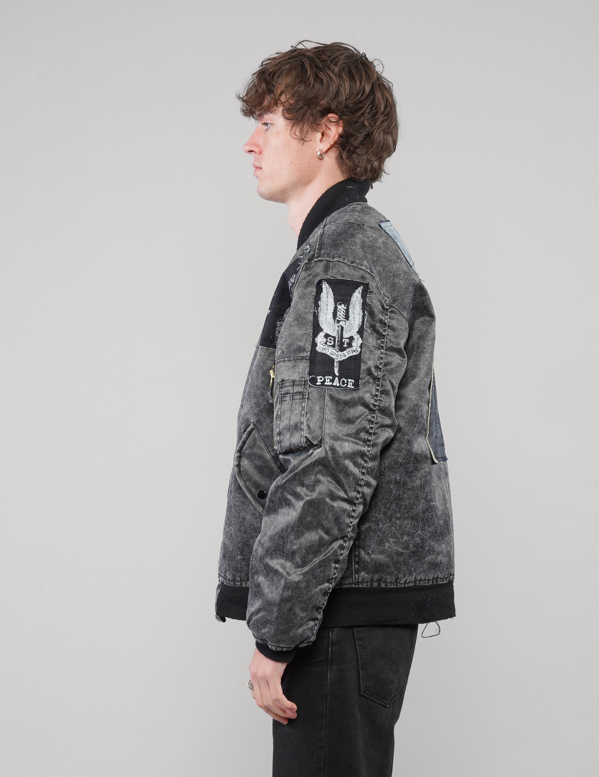 Left-side view of a model in the charcoal bomber jacket, showing the Peace sleeve patch and black ribbed trims.