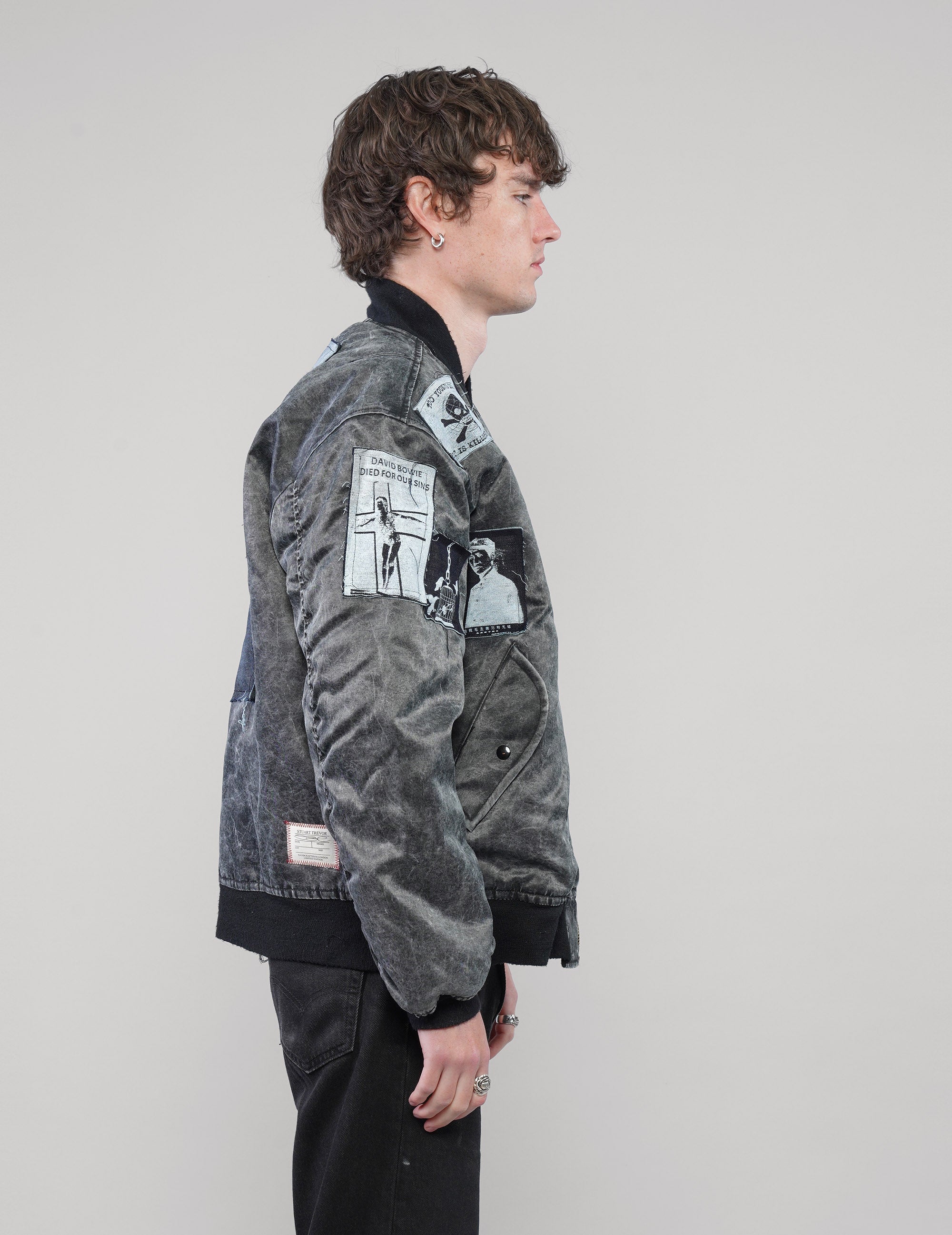 Right-side view of a model wearing the charcoal bomber jacket, showing graphic sleeve patches and ribbed cuffs.